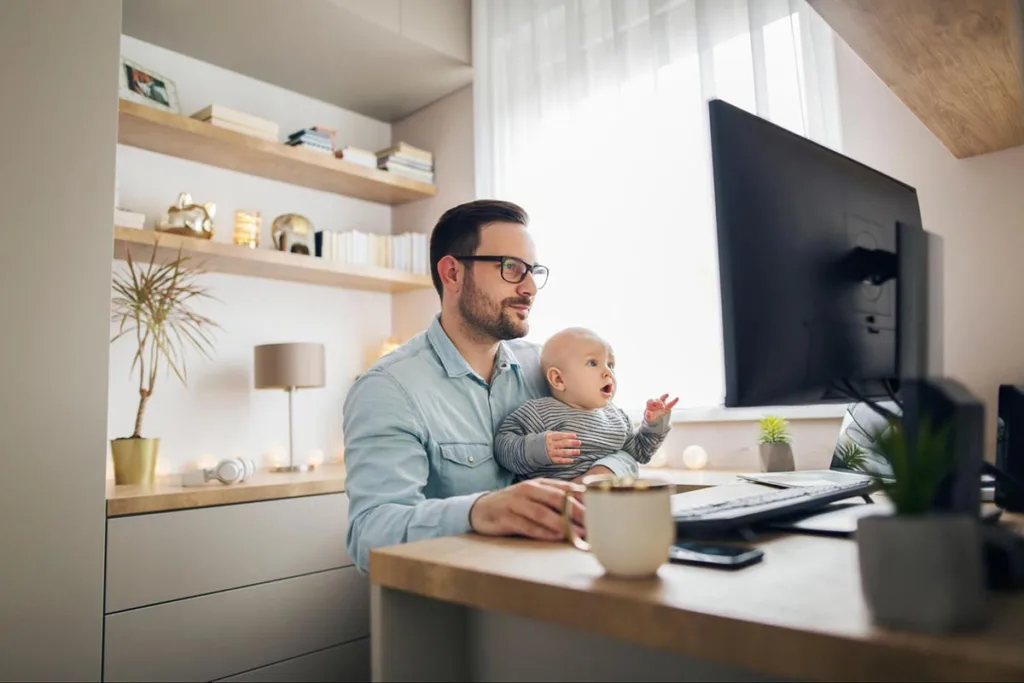 Entrepreneur holding his baby and working with laptop while working from home