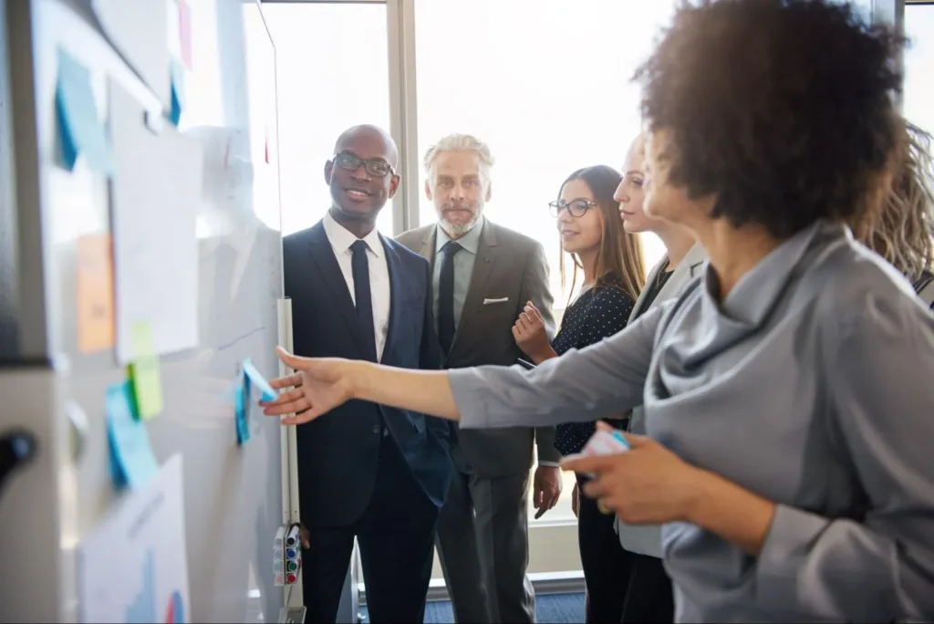 A group of marketing professionals around a white board collaborating while one woman grabs a post-it note