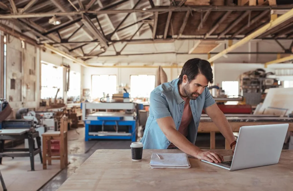 Business owner in a furniture making workshop exploring free advertising options on a laptop