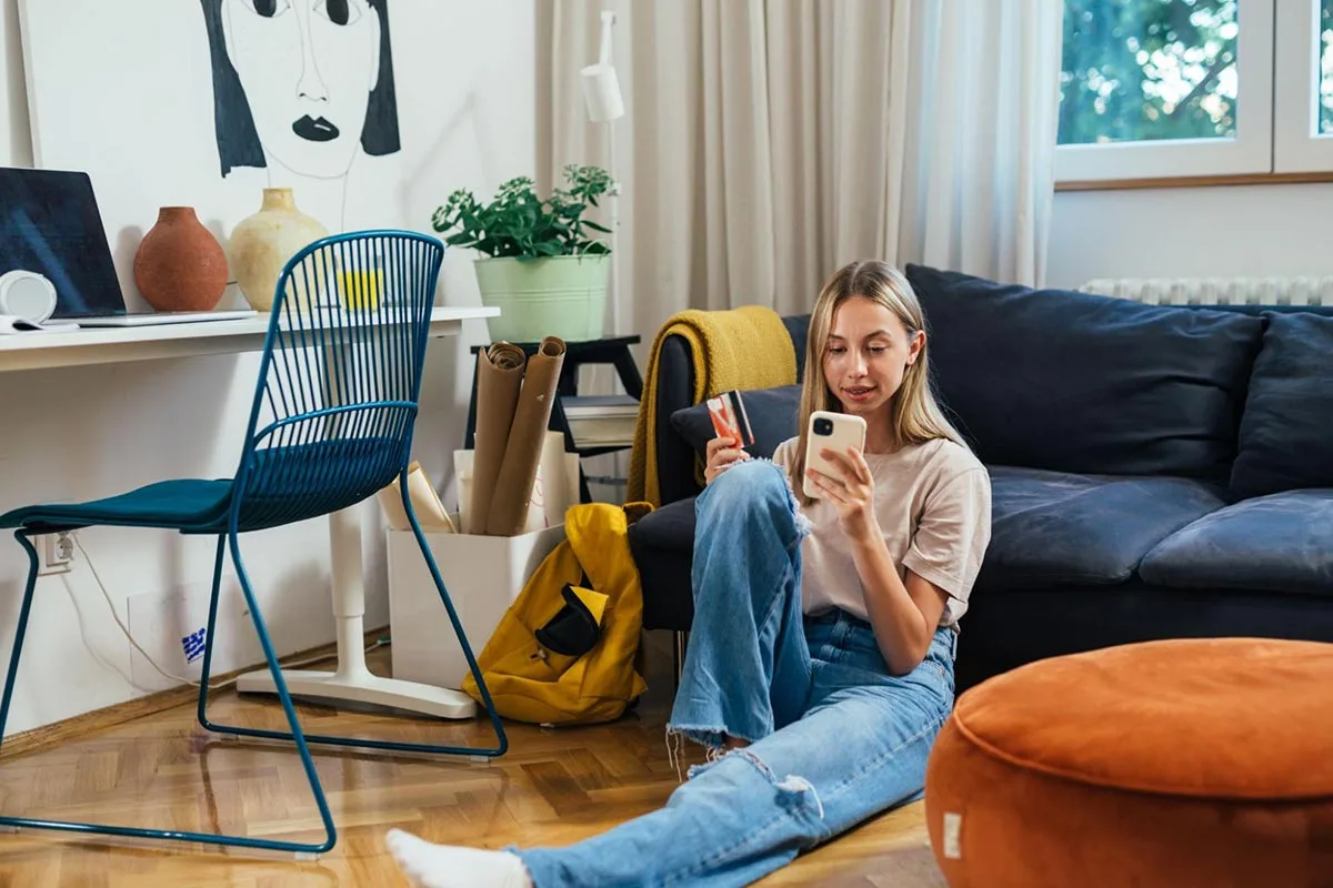 Young woman using bank card to pay on her phone while sitting on her living room floor