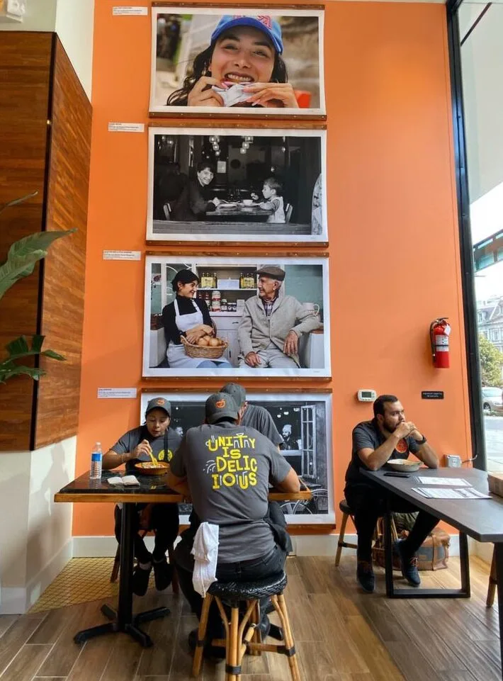 Photo of Immigrant Food servers eating a meal at the restaurant, under framed photos of smiling people on the wall