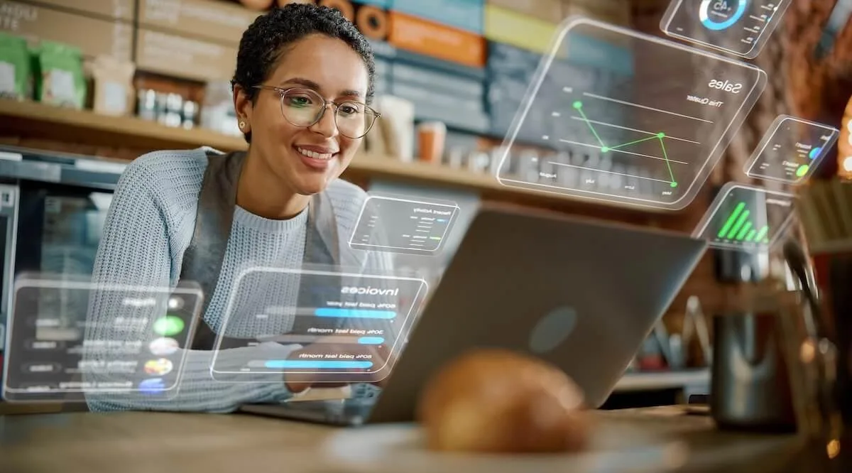 Smiling cafe owner using laptop to review table management software with futuristic digital interface overlays.
