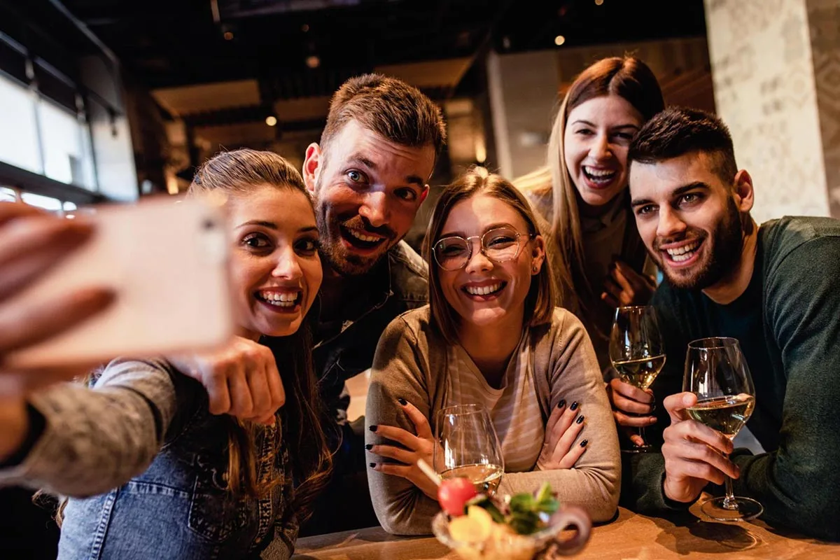 Group of friends toasting drinks and taking a selfie at a restaurant table.