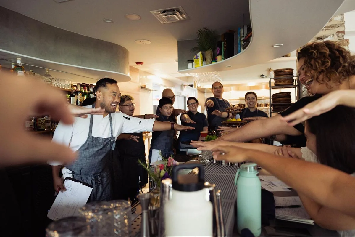Photo of Kaya Executive Chef Lordfer Lalicon and co-owner Jamilyn Salonga Bailey leading their team in a pre-dinner huddle, with the whole group putting their hands in the middle to cheer, from Lee Romero.
