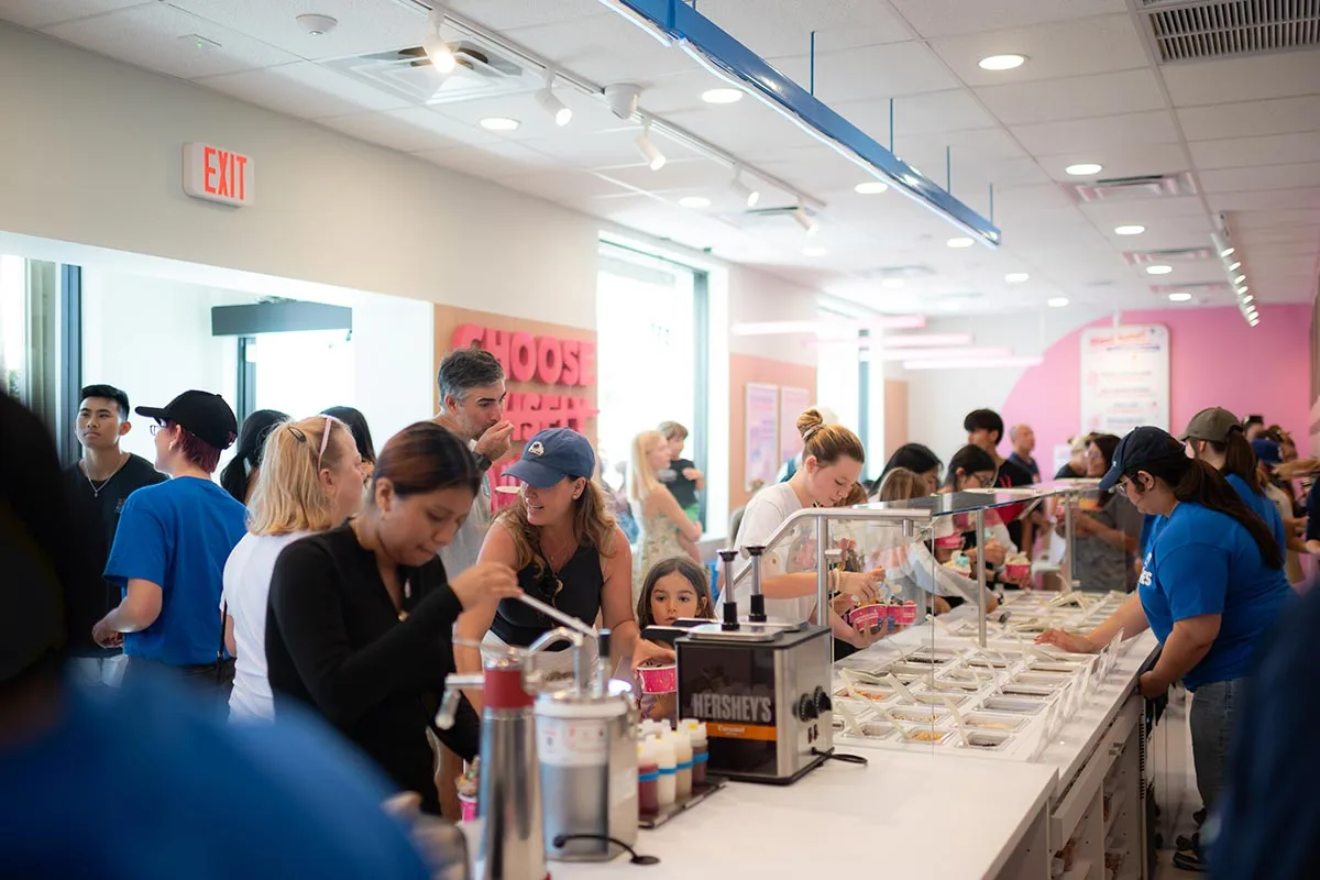 Photo of a large group of customers helping themselves to the self-serve topping bar inside a 16 Handles store.