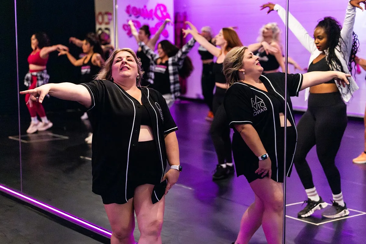 Photo of DivaDance CEO Jami Stigliano giving direction during a class while dancers move in the background.
