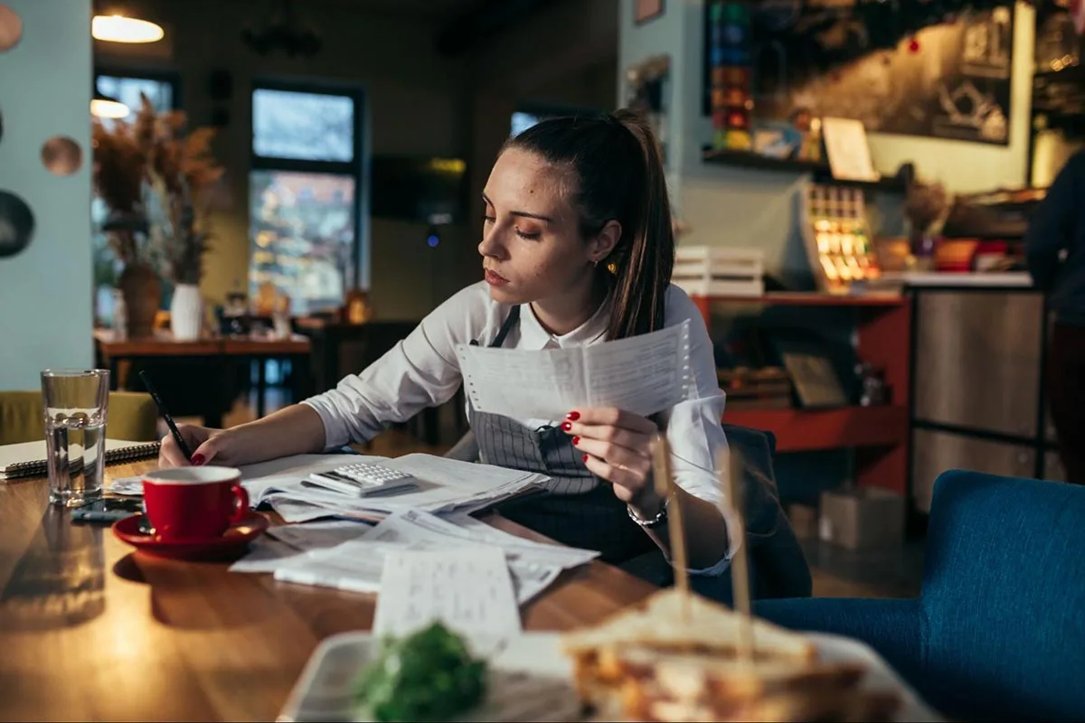 A restaurant manager is calculating expenses at a table with documents and a calculator.