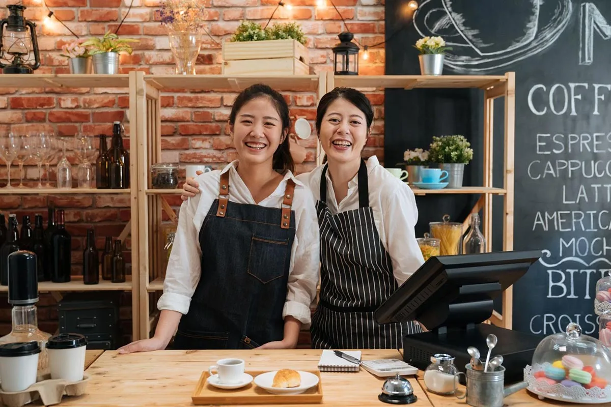 Two baristas are smiling together inside a coffee shop.