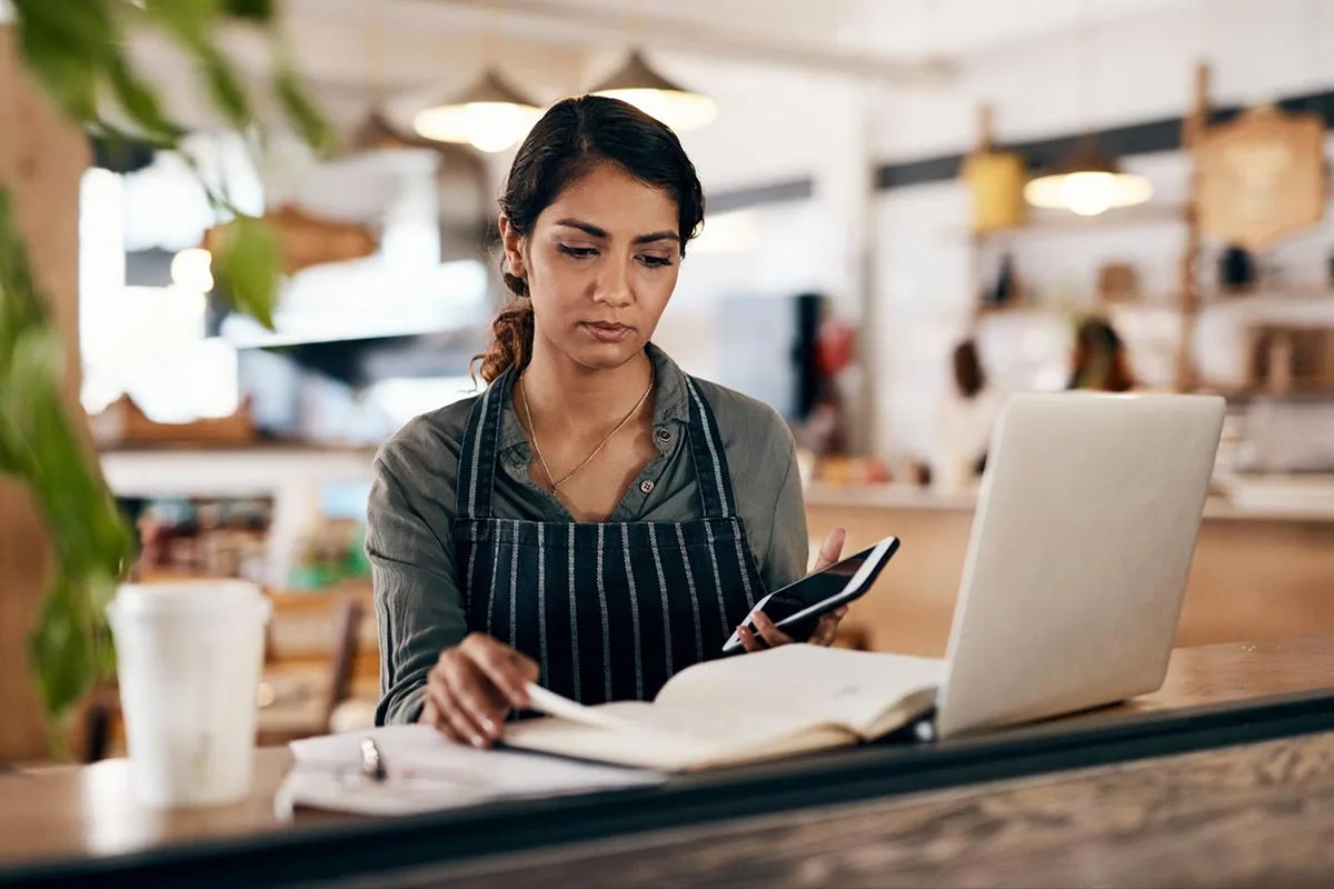 A restaurant manager is holding a phone while using a laptop.